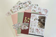 A flat lay of floral scrapbooking paper sheets and sticker sheets arranged on a pale yellow background. The sheets feature pink and cream roses, green leaves, vintage-style stamps, buttons, and a rustic wood texture.